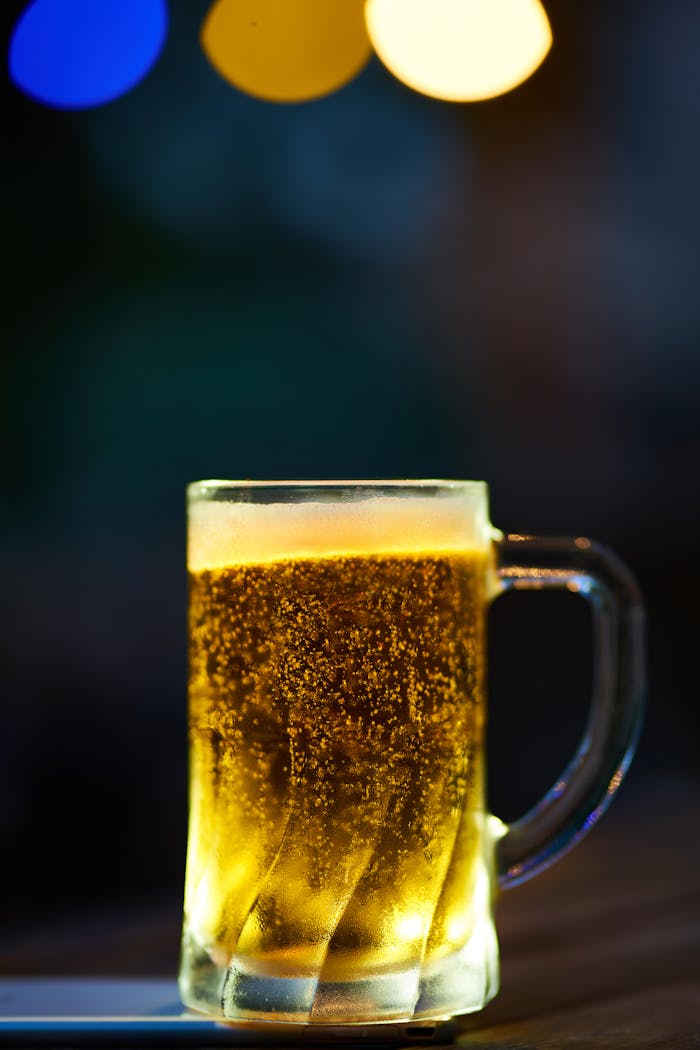 A glass mug of cold beer with frothy head and bokeh lighting effect in a dim setting.